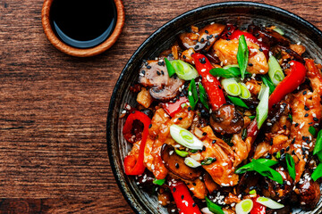 Stir fry chicken with paprika, mushrooms and chives in bowl. Asian cuisine dish. Wooden kitchen table background, top view