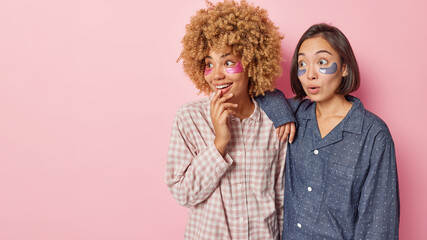Two women of different race apply hydrogel patches under eyes undergoes skin care procedures try to reduce puffiness dressed in nightwear isolated over pink background blank space for your promo