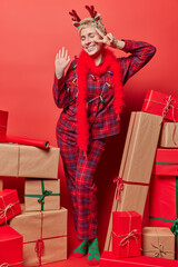 Joyful short haired woman prepares for domestic party on Christmas makes peace sign over eye smiles gladfully dressed in nightwear prepares presents for her friends red background. Winter holidays