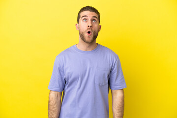 Handsome blonde man over isolated yellow background looking up and with surprised expression