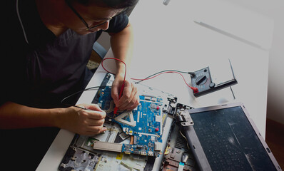 young man wearing glasses who is a technician computer technician A laptop motherboard repairman is using an IC meter to find faults on the motherboard for repair on his workbench.