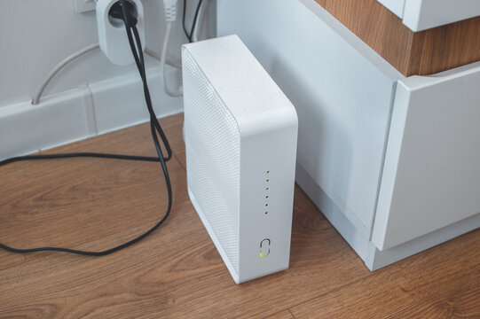 A modern Wi-Fi router plugged into an outlet, white and without antennas, stands on the floor in a home room.