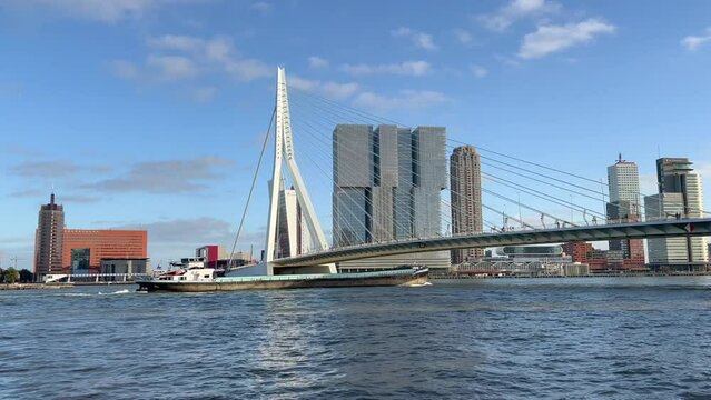 Cargo, freight ship is going under Erasmus bridge (Erasmusbrug). Rotterdam city skyline with skyscrapers in the background. Motion of river waves. People and cars on bridge. Rotterdam, Netherlands