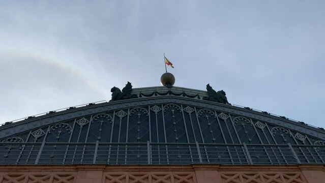 Spanish flag is waiving on top of Madrid Atocha train, railway station building roof. Main, front entrance. Bright blue sky. Madrid, Spain