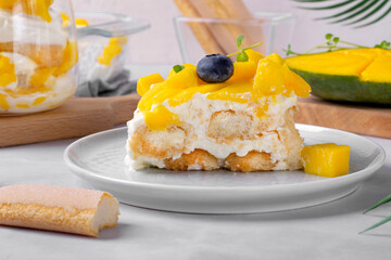 Piece of tiramisu cake with mango topped with fruit slices served on the plate on white table. Italian dessert variation