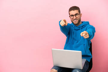 Young man sitting on a chair with laptop points finger at you while smiling