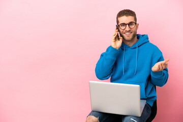 Young man sitting on a chair with laptop keeping a conversation with the mobile phone with someone