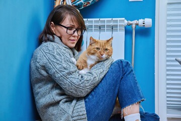 Woman in knitted woolen sweater with pet cat warming near heating radiator