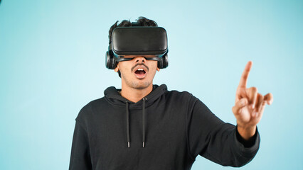 Young Indian Asian man using VR headset, glasses or goggles, isolated over blue colour indoor studio background, shocked surprised or amazed concept