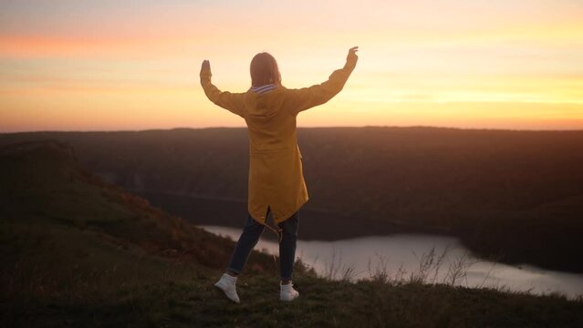 Camera follows young woman in yellow jacket running up on top of mountain summit at sunset, jumps on top of rocks, raises arms into air, happy and drunk on life, youth and happiness