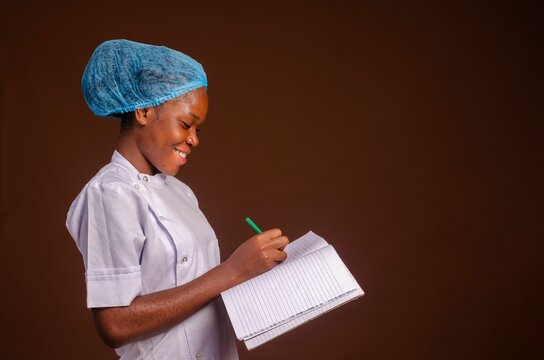 Smiling Nigerian female medic writing on a book isolated on a brown background