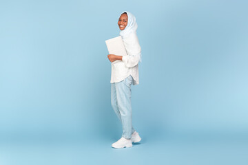 Happy african american woman in hijab holding laptop and walking on blue studio background, full...