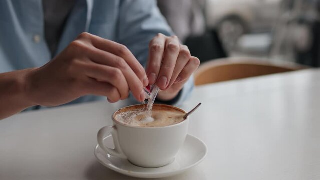 Woman's hands put sugar packaging in ready cup of cappuccino, Sugar sticker in cappuccino cup, Woman sits in coffee shop ordered a cup of coffee with sugar, having a lunch break in a restaurant
