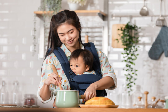 Happy New Asian Mom Hold And Carry Baby On Baby Bag In Front And Cooking Breakfast In The Morning. Smile Mother Carrying Little Baby Infant Preparing Healthy Food.Mom And Baby Spending Time At Kitchen