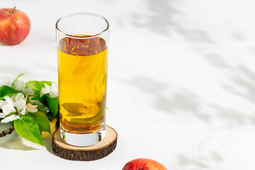 Apple juice. Full glass of natural apple juice on a wooden podium on a white table with red apples, blossom branches, shadows and copy space. Summer natural drink. Vegan and vegetarian healthy food