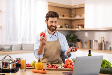 Happy mature caucasian male with beard in apron has video call, makes video holds tomato with hands