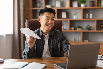 Smiling middle aged asian male making airplane with paper, planning strategy at table with laptop