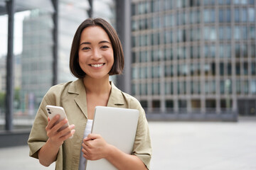 Portrait of smiling asian girl with laptop, holds mobile phone and looks happy at camera, stands on...