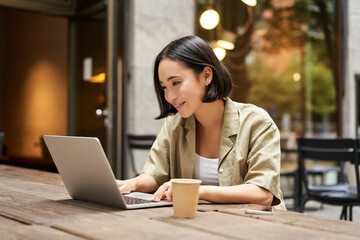 Young asian woman, digital nomad working remotely from a cafe, drinking coffee and using laptop, smiling - obrazy, fototapety, plakaty
