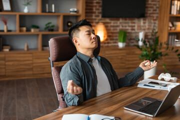 Obraz premium Relaxed adult asian man with closed eyes meditation, rests at table with laptop, enjoys silence and comfort
