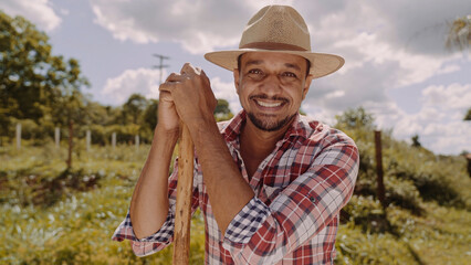 Portrait of young man in the casual shirt holding his hoe in the farm. Farm tool. Latin man. - obrazy, fototapety, plakaty