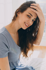 Vertical indoor image of lovely adorable charming beautiful and pretty beautiful brunette female sitting on bed against window, touching her forehead, having long wavy hair, dressed in gray