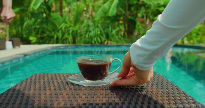 Waiter woman hand serving americano coffee by the swimming pool. Outdoor breakfast on holiday. Asian girl brings cup of caffein drink on table in tropical resort. Travel weekend vacation concept.