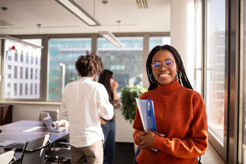 Black girl in office in foreground is looking at camera - obrazy, fototapety, plakaty