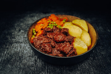 Hearty goulash with peas and carrots and boiled potatoes in a bowl with a dark background