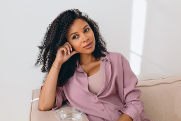 Fototapeta na wymiar Horizontal indoor image of petty charming african american female with gorgeous long curly hair, leaning head on hand sitting on sofa, looking aside with positive thoughtful facial expression