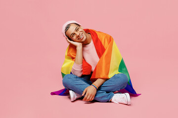 Full body minded happy cheerful young gay man wear sweatshirt hat wrapped in flag sitting on floor isolated on plain pastel light pink color background studio portrait. Lifestyle lgbtq pride concept.