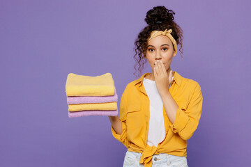 Young housekeeper woman wears yellow shirt tidy up hold in hand pile of clothes t-shirts cover mouth with arms looking camera isolated on plain pastel light purple background studio Housework concept