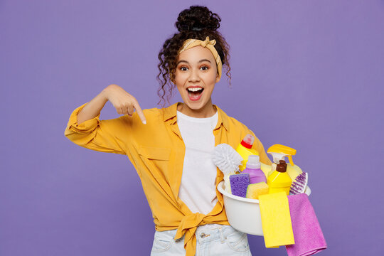 Young surprised fun housekeeper woman wear yellow shirt while doing tidy up point finger down on basin with detergent bottles isolated on plain pastel light purple background studio Housework concept.