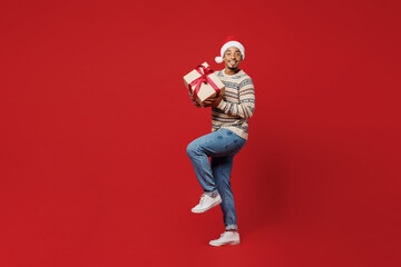 Side view full body fun merry young man wear Christmas sweater Santa hat posing hold present box with gift ribbon bow raise up leg isolated on plain red background Happy New Year 2023 holiday concept