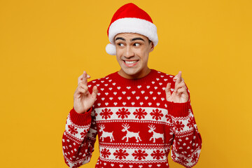 Merry young man wear red knitted christmas sweater Santa hat posing wait special moment keeping fingers crossed, making wish hope trust isolated on plain yellow background Happy New Year 2023 concept