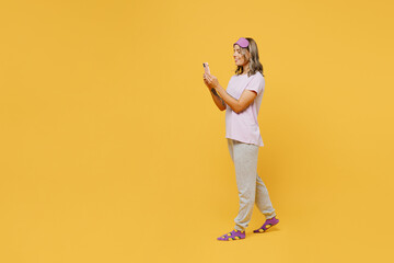 Fototapeta na wymiar Full body side view young woman wears purple pyjamas jam sleep eye mask rest relax at home hold in hand use mobile cell phone go isolated on plain yellow background studio portrait. Night nap concept.