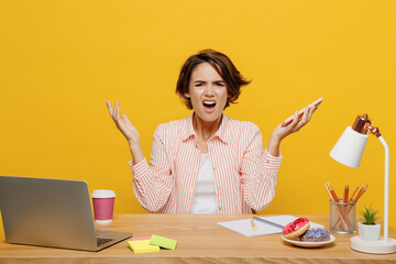 Young indignant annoyed sad employee business woman wear casual shirt sit work at office desk hold mobile cell phone spread hands isolated on plain yellow color background. Achievement career concept.