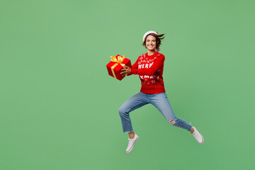 Full body merry young woman wear knitted xmas sweater Santa hat posing hold gift present box jump high isolated on plain pastel light green background. Happy New Year 2023 celebration holiday concept.