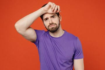 Fototapeta na wymiar Young exhausted tired sad brunet caucasian man 30s wear casual basic purple t-shirt hand on forehead suffer from headahce isolated on plain orange background studio portrait. People lifestyle concept.