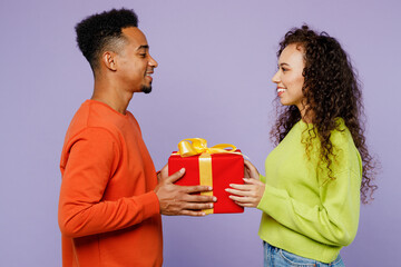 Side view young couple two friend family man woman of African American ethnicity in casual clothes together give red present box with gift ribbon bow isolated on pastel plain light purple background