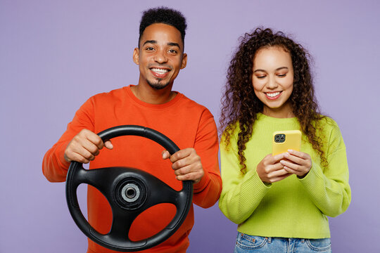 Young couple two friends family man woman of African American ethnicity wear casual clothes together hold steering wheel driving car use mobile cell phone isolated on pastel plain purple background.