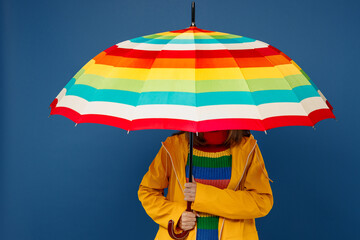 Young woman wear sweater yellow waterproof raincoat outerwear hold cover face with opened colorful umbrella isolated on plain dark royal navy blue background Outdoors wet fall weather season concept. - obrazy, fototapety, plakaty