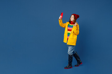 Full body young woman wear sweater red hat yellow waterproof raincoat do selfie shot on mobile cell...