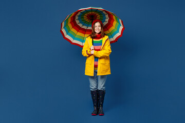 Full body young woman in sweater red hat yellow waterproof raincoat outerwear hold opened colorful umbrella isolated on plain dark royal navy blue background Outdoors wet fall weather season concept - obrazy, fototapety, plakaty