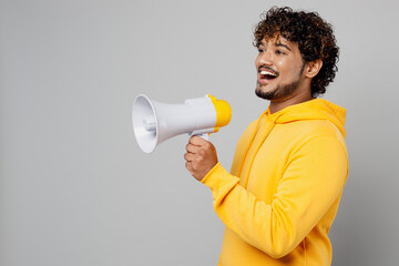 Side view young Indian man 20s he wearing casual yellow hoody hold scream in megaphone announces discounts sale Hurry up isolated on plain grey background studio portrait. People lifestyle portrait.