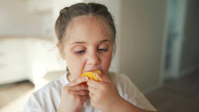 Little girl at kitchen table. Child eats yellow oranges. Happy child eats fruits. Vegetarian girl in kitchen.Concept of proper nutrition.Girl eats yellow fruit oranges.Juicy fruits proper healthy diet