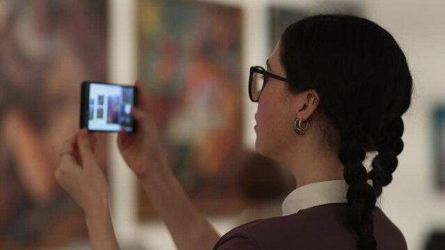 Side view of young pretty Caucasian woman wearing eyeglasses take photo with smartphone in art gallery. Visiting excursion in museum. Concept of educational and cultural tourism