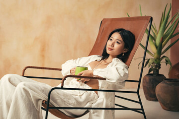 Asian woman lying on lounger on spa resort, holding cup of coffee in hands