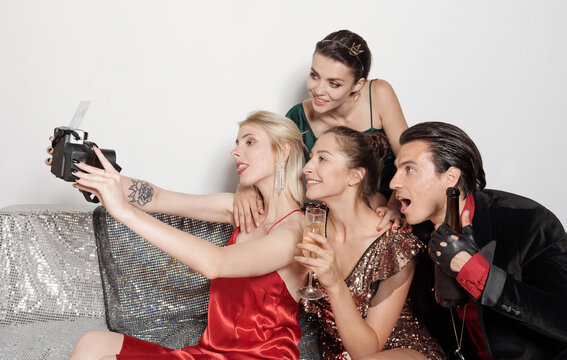 Three girls and one guy sitting together on sofa with drinks taking group selfie at New Year party 