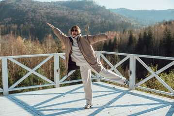 a girl in warm clothes stands on the terrace against the background of mountains;
jumps and rejoices in sunny weather